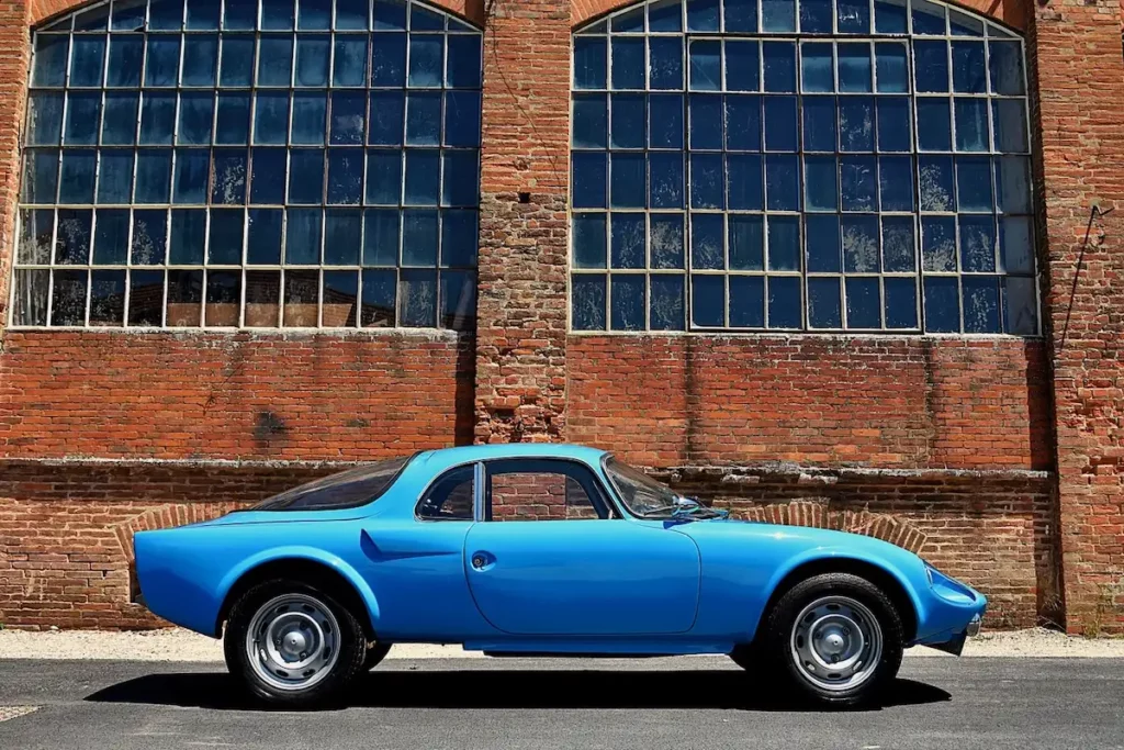 Voiture de collection bleue Alpine Djet stationnée devant un bâtiment en briques rouges aux grandes fenêtres vitrées – photographie artistique en lumière naturelle.